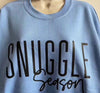 Blue cotton crewneck sweatshirt with "Snuggle Season" text, ideal for warmth and comfort in colder months.