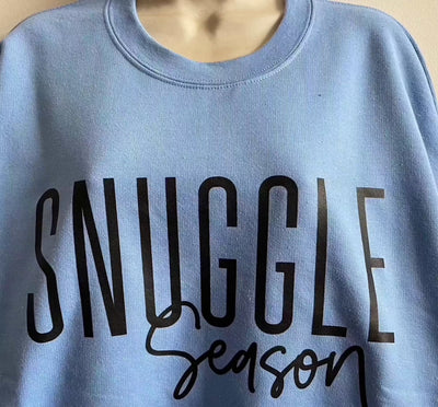 Blue cotton crewneck sweatshirt with "Snuggle Season" text, ideal for warmth and comfort in colder months.