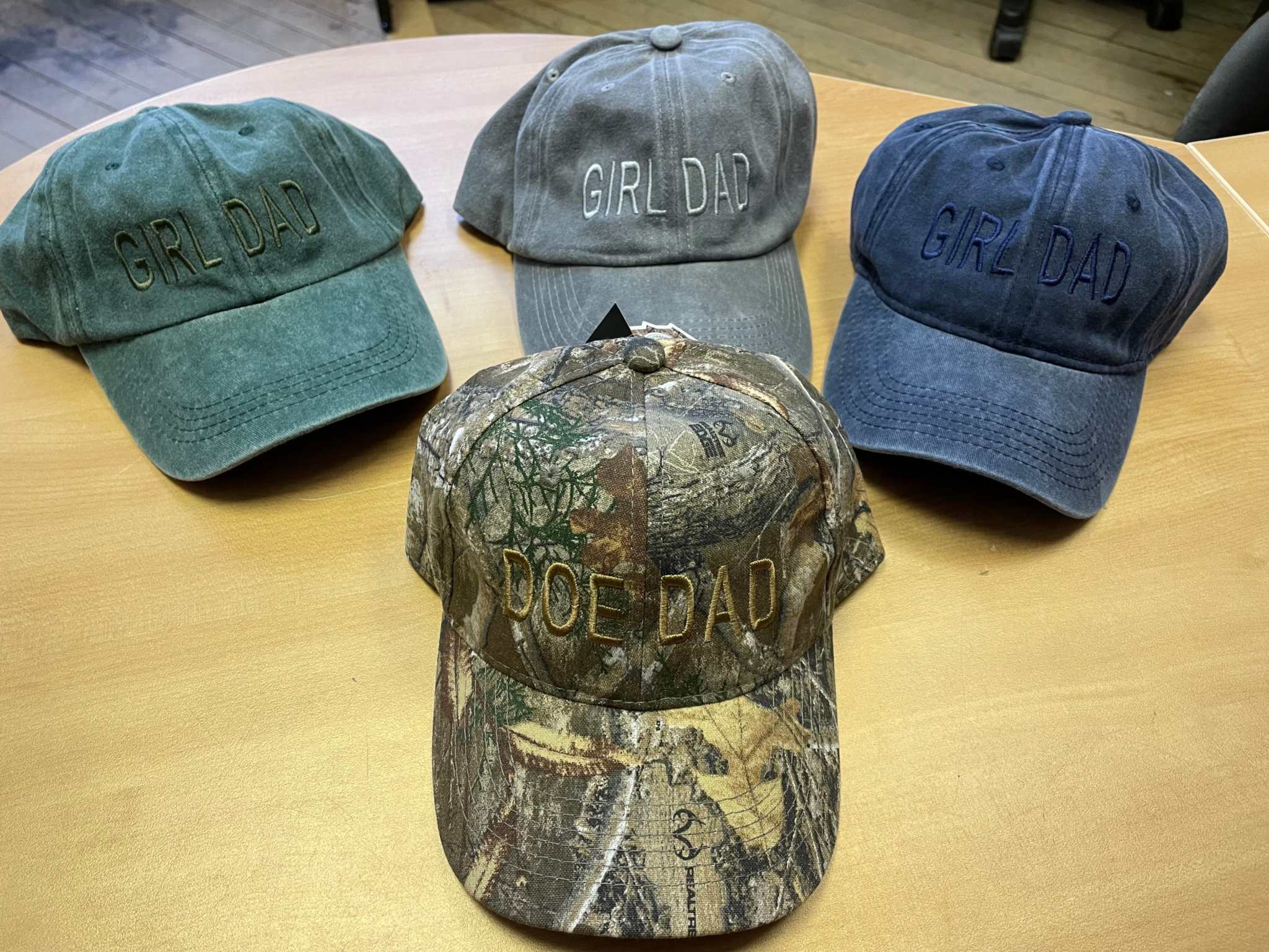 Four adjustable baseball hats on wooden table, three with "GIRL DAD" text in green, gray, and blue, one camo cap with "DOE DAD" text, cotton brushed fabric style