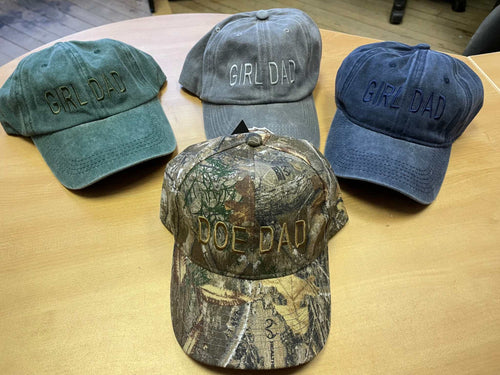 Four adjustable baseball hats on wooden table, three with "GIRL DAD" text in green, gray, and blue, one camo cap with "DOE DAD" text, cotton brushed fabric style