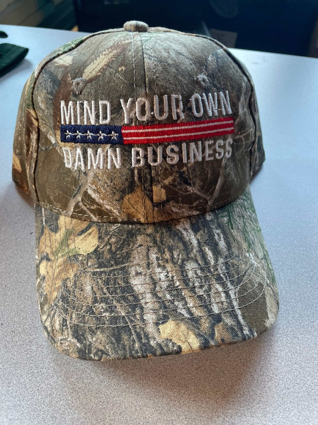 Camo hat with "Mind Your Own Damn Business" embroidered slogan and American flag design