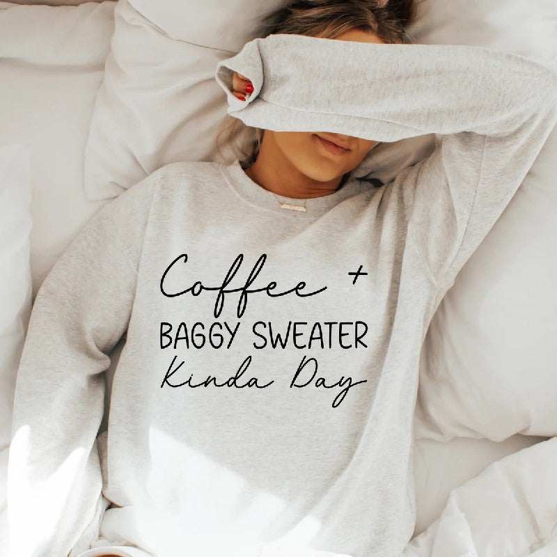 Woman wearing gray Coffee + Baggy Sweater Kinda Day sweatshirt relaxing on white bed