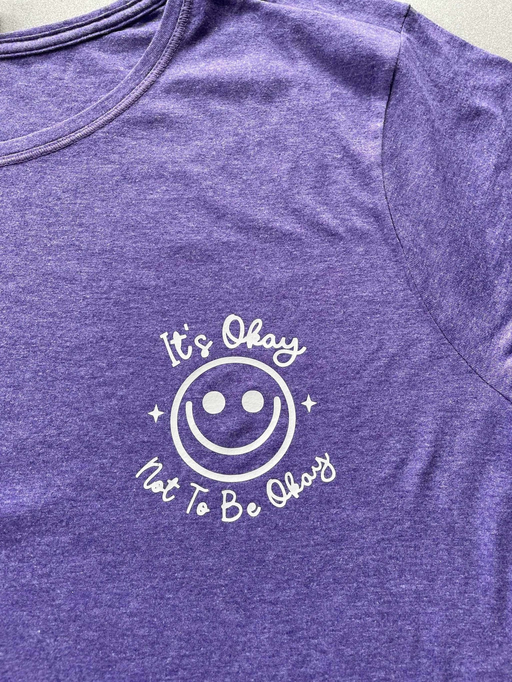 Purple heather women's graphic tee with "It's Okay Not to Be Okay" smiley design