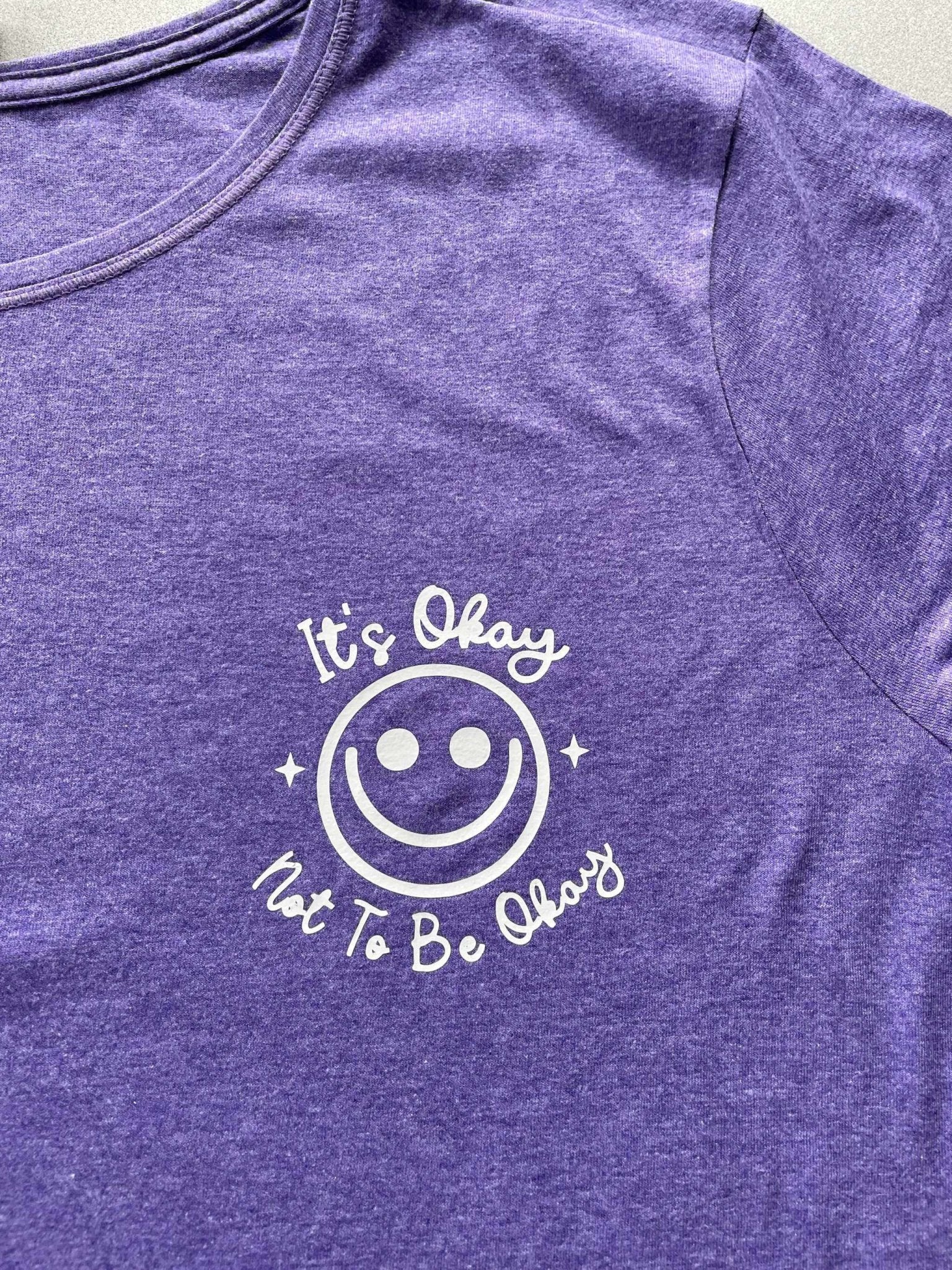 Purple heather women's graphic tee with "It's Okay Not to Be Okay" smiley design