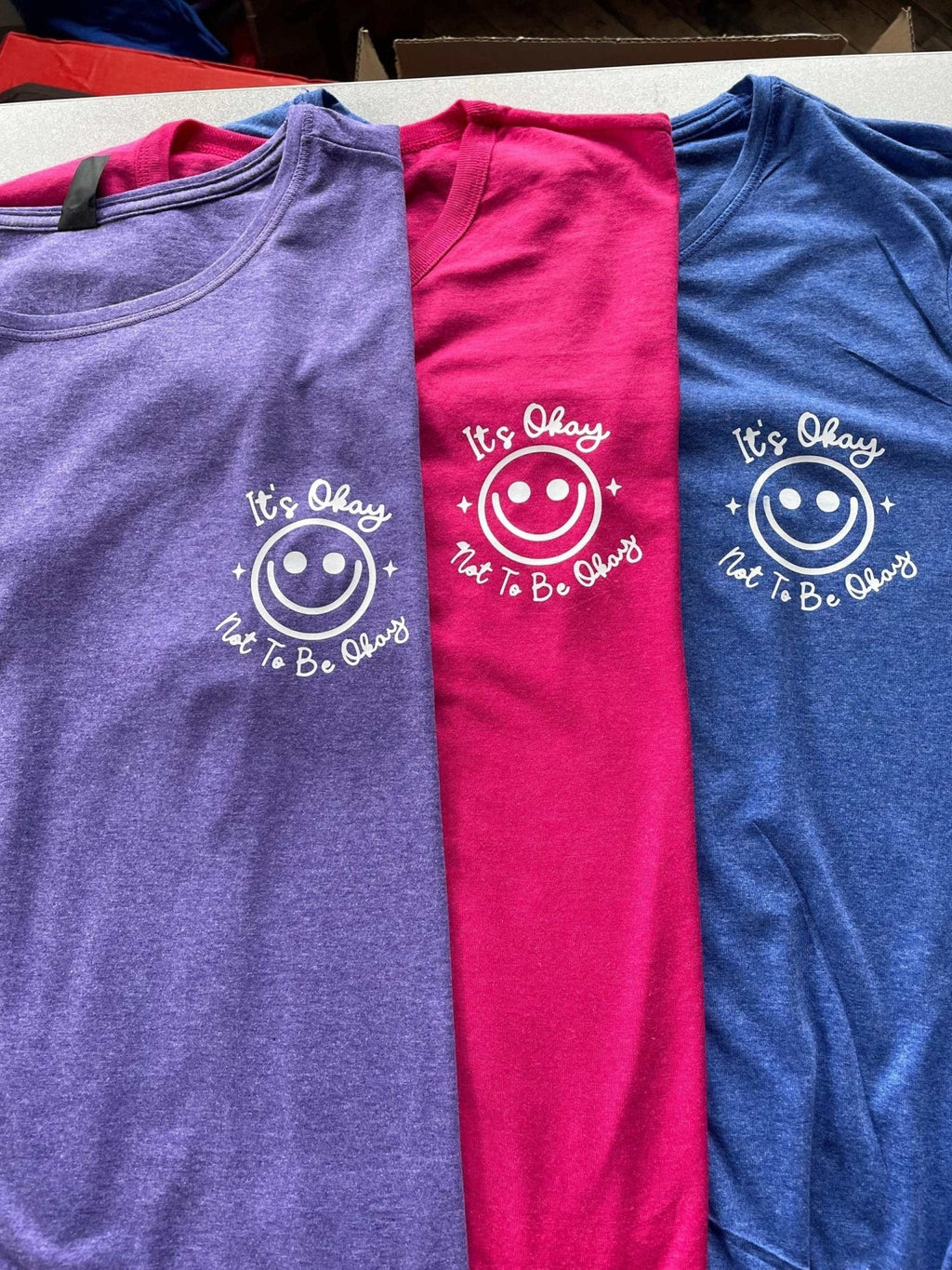 Women's graphic tee shirts in purple, red, and blue heather with "It's Okay Not To Be Okay" smiley design