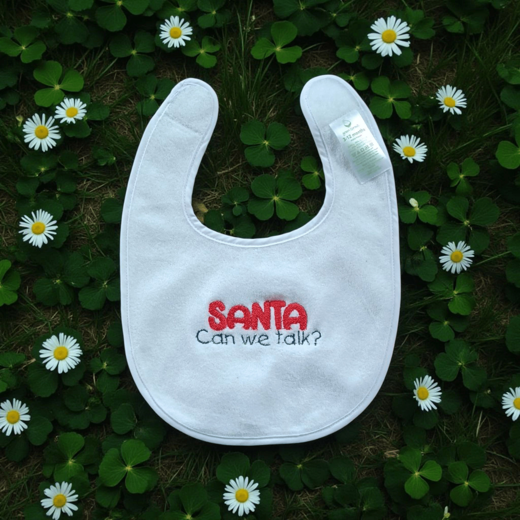 White holiday baby bib with "Santa Can We Talk?" embroidery on a grass and flower background