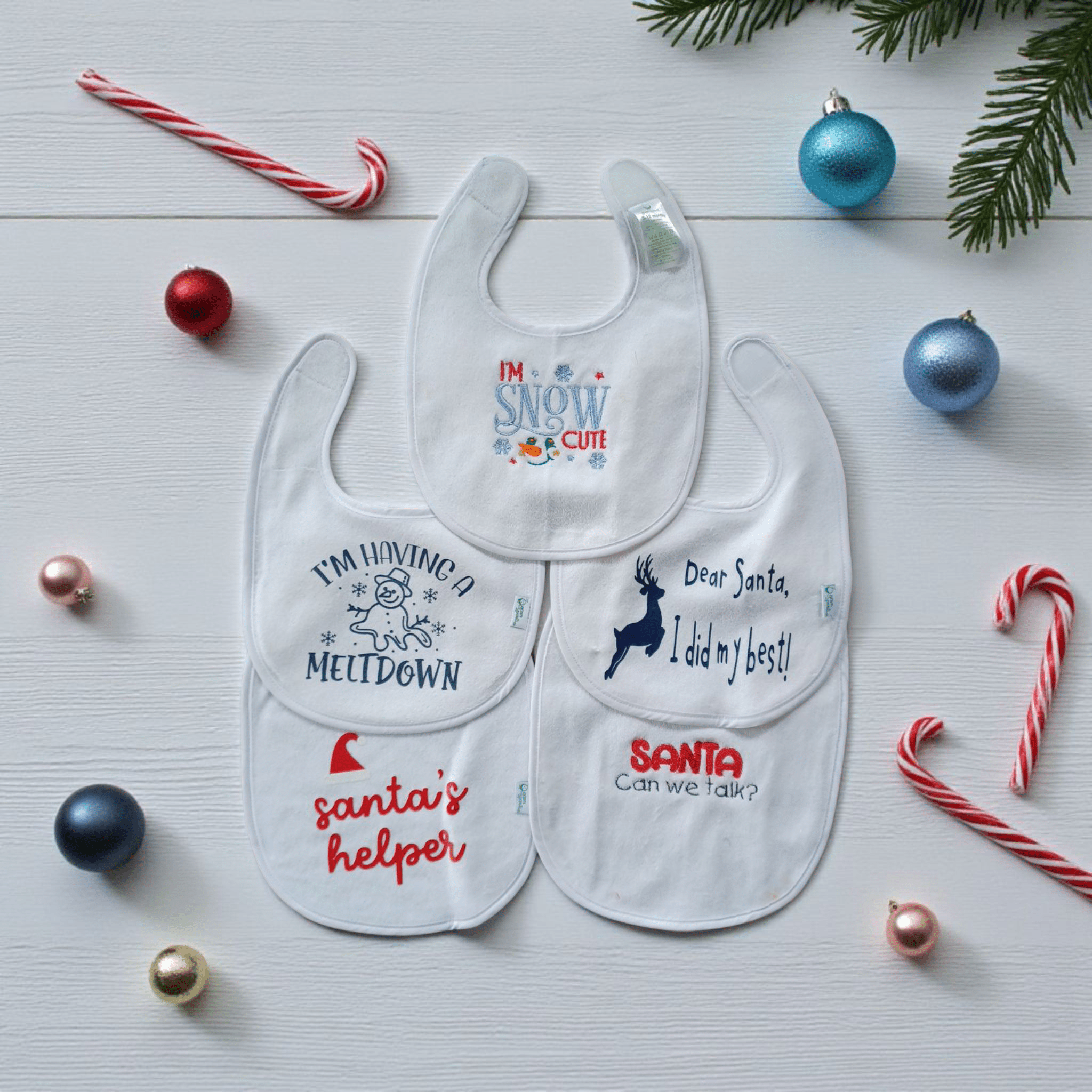 Set of four white Holiday Baby Bibs with festive embroidery and prints, surrounded by Christmas decorations and candy canes on a wooden background.