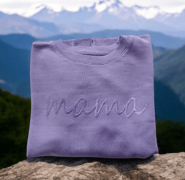 Folded lavender cotton sweater with embroidered word Mama displayed outdoors with mountains background