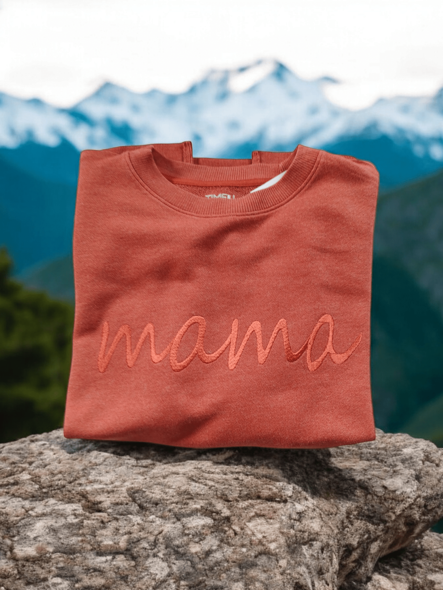 Folded rust orange cotton sweater with embroidered "Mama" text on a rock outdoors